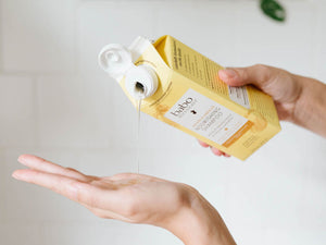 A person holds a yellow Babo Hair Growth & Scalp Care carton while pouring a clear gel into their palm against a light neutral background.
