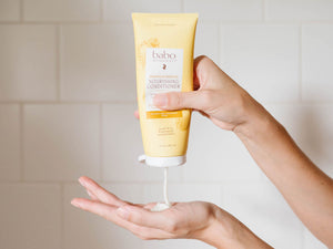 A person's hands squeezing a yellow tube of baboo shine Nourishing Conditioner, with white conditioner dripping onto another hand against a light tiled wall background.