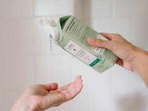 A person's hands pour a clear, viscous liquid from a light green carton labeled 'bioderma' onto an open palm, set against a soft-focus white tiled background.