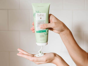 A hand holds a tube of babo haircare Volumizing Conditioner above another hand dispensing white conditioner cream, set against a soft beige tiled background.