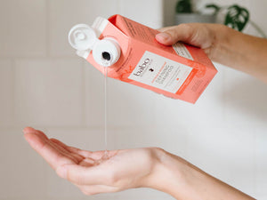 A person holds an orange-brown carton of babko Vitamin S serum and pours a thin stream of the clear serum into their open palm, set against a soft, light beige tiled background.
