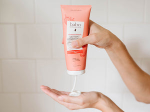 A hand holds a squeeze tube of baboo botanicals Ragadeep Defining Conditioner, with white cream being dispensed into another hand against a white tiled background.