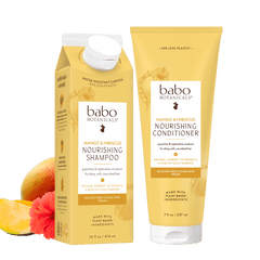 Babo Botanicals haircare set featuring a carton of nourishing shampoo and a tube of nourishing conditioner, both in a warm yellow-gold color, shown on a white background with citrus and flower props.