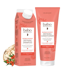 Babo Botanicals hair care set featuring a water-resistant carton of Defining Shampoo and a tube of Defining Conditioner in a coral pink color, shown with a small ingredient graphic of oats and red chilis on a white background.