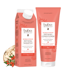 Babo Botanicals hair care set featuring a water-resistant carton of Defining Shampoo and a tube of Defining Conditioner in a coral pink color, shown with a small ingredient graphic of oats and red chilis on a white background.