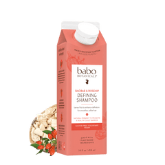 A carton of Babo Botanicals Defining Shampoo in a coral pink color, shown with a white cap and styled with dried botanical ingredients in a small bowl on a white background.