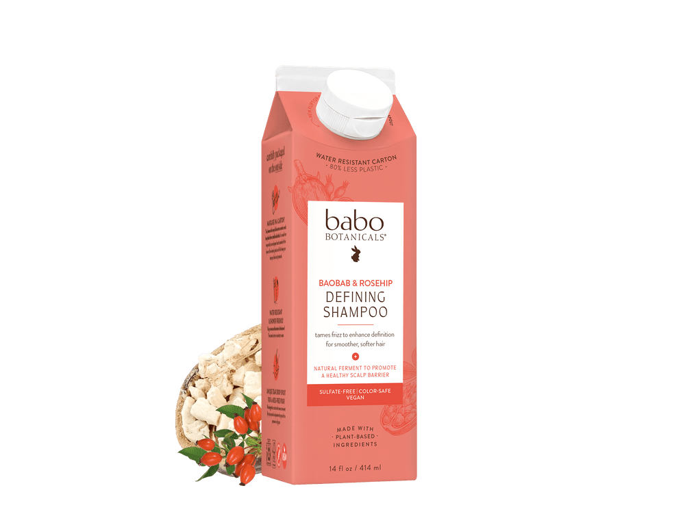 A carton of Babo Botanicals Defining Shampoo in a coral pink color, shown with a white cap and styled with dried botanical ingredients in a small bowl on a white background.