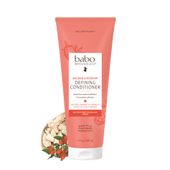 A squeeze tube of babo Botanicals Barba & Beardshop Defining Conditioner in a coral pink color, shown with a small decorative graphic of a beard balm bowl at the bottom left, on a solid white background.